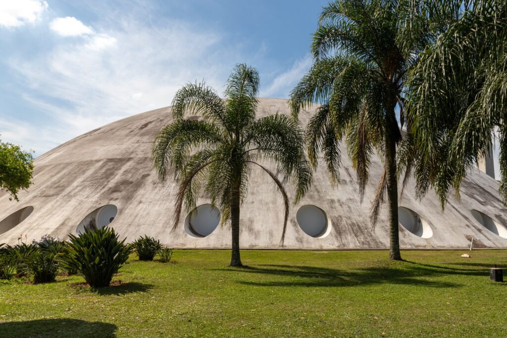 Vista externa da Oca, pavilhão em forma de cúpula branca projetado por Oscar Niemeyer no Parque Ibirapuera, com palmeiras em primeiro plano